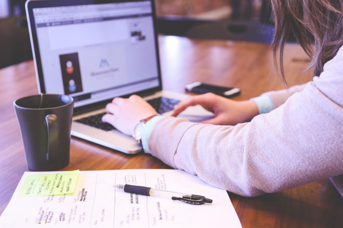 Person using a laptop with a coffe mug and notepad beside them 