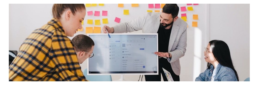 A man holding up a picture showing a screenshot of a computer screen, in front of three people in a meeting