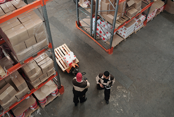 Two men standing in a warehouse, with racking either side of them that have boxes on the shelves.