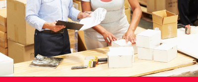 One person packing items in boxes whilst the other is reading from paper attached to a clipboard