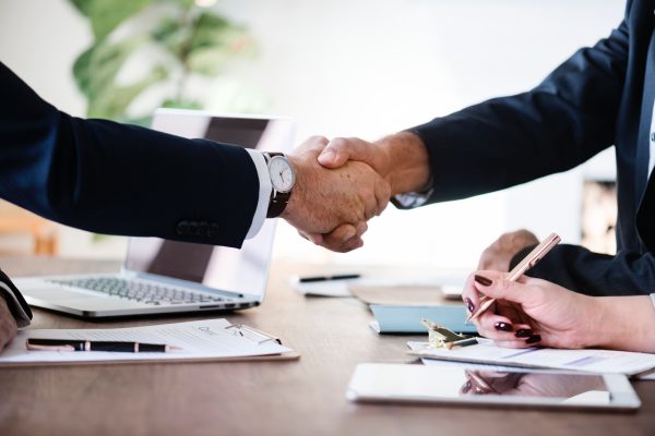 Two people wearing suits shaking hands across a table.